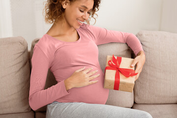 Baby shower gift. Black pregnant woman holding gift box, resting on sofa