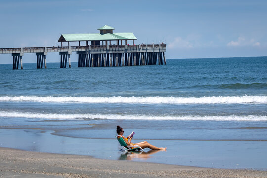 Beautiful young woman sits alone in beach chair along the rolling waves reading a book in bikini with Folly Beach pier in background