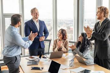 A group of four business professionals are gathered around a conference table, clapping and smiling...