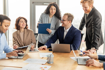 A diverse group of business professionals gathered around a table in a modern office, collaborating on a project. Two people are focused on their laptops, while another holds a clipboard with papers