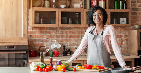 Attractive black woman cooking healthy veggies salad at kitchen, smiling at camera, copy space