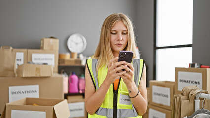 Blonde woman volunteer texting indoors surrounded by donation boxes