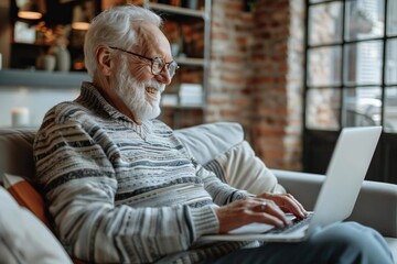Obraz premium Happy senior man with glasses sitting on a couch, smiling and laughing while using a laptop, looking at the screen.