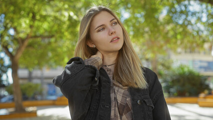 A young caucasian woman with blonde hair outdoors, surrounded by nature, looking pensive in a park setting.