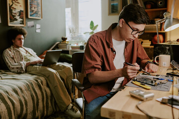 Young technician soldering small computer details while his friend working remotely on bed in...
