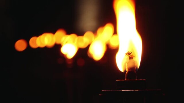 A close up shot of a line of lit torches, their flames glowing warmly in the dark night. The foreground torch is in sharp focus, while the others blur into the background, creating a serene
