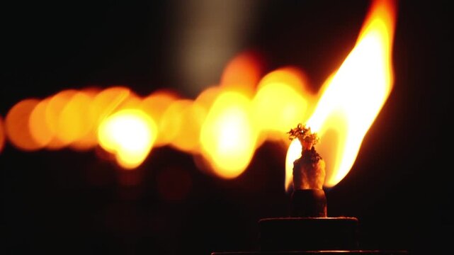 A close up shot of a line of lit torches, their flames glowing warmly in the dark night. The foreground torch is in sharp focus, while the others blur into the background, creating a serene