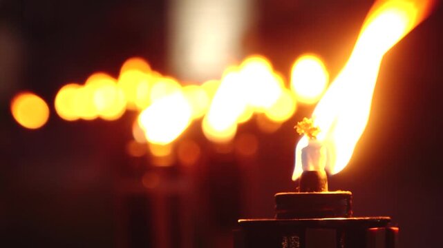 A close up shot of a line of lit torches, their flames glowing warmly in the dark night. The foreground torch is in sharp focus, while the others blur into the background, creating a serene