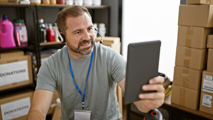 Mature hispanic man with headset using tablet in warehouse full of donations and supplies.