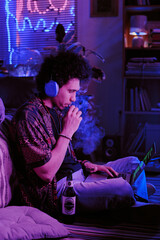 Curly-haired developer taking inhalation of sweet vape while checking codes on laptop