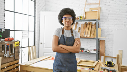 Confident black woman with curly hair wearing safety glasses stands arms crossed in an indoor...
