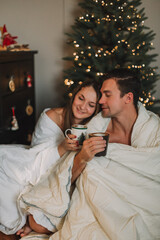 A couple of lovers wrapped in a white blanket hugging under a Christmas tree. Husband and wife drink cocoa. Romantic morning under the Christmas tree