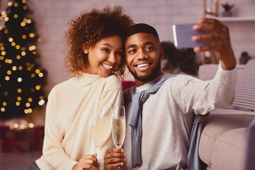 Christmas time. Afro couple taking selfie against xmas tree, drinking champagne