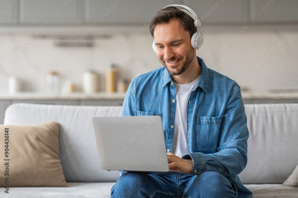 Aufkleber a man in a denim shirt sits on a couch in a living room. he is using a laptop computer and smiling. 