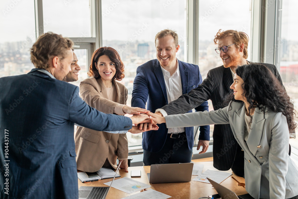 Fototapete A group of five diverse business professionals stand around a conference table, smiling and stacking their hands on top of each other in a gesture of unity and teamwork.