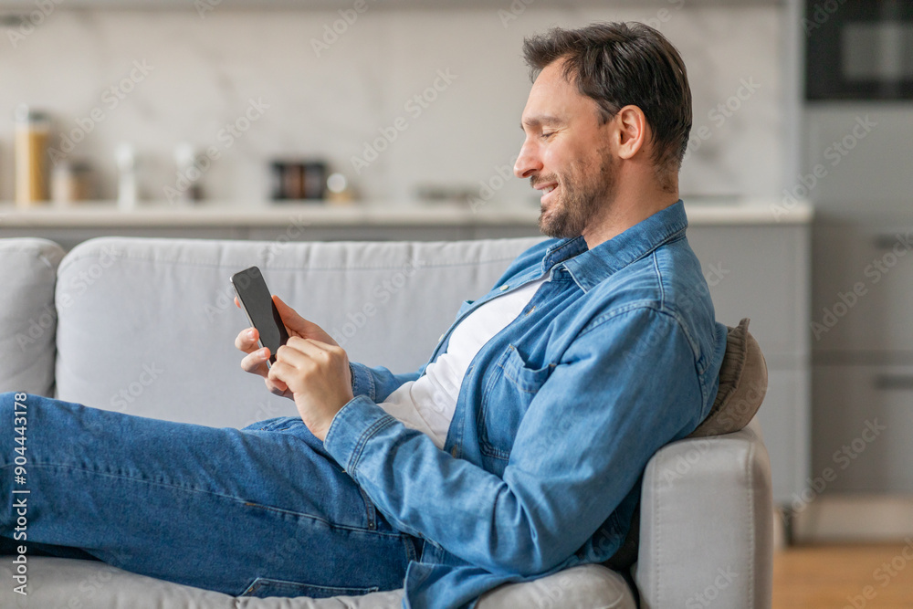 Poster A man is sitting on a couch in a living room while using a smartphone. He is smiling as he looks at the phone. He is wearing jeans and a blue denim shirt.