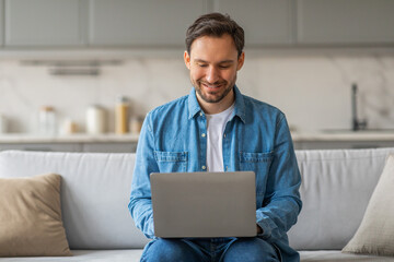 A man wearing a blue denim shirt and jeans sits on a white couch in a modern home, using a laptop...