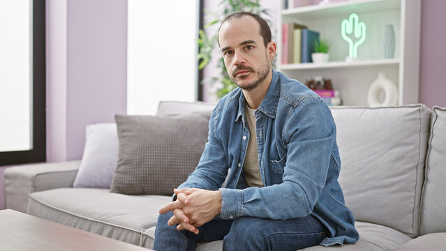 Hispanic bald man with beard in denim sitting thoughtfully on a gray sofa indoors. - Powered by Adobe