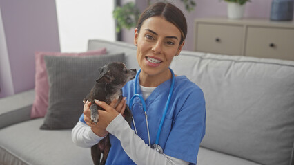 Fototapeta premium A smiling woman in scrubs holding a chihuahua in a cozy living room setting, suggesting a veterinarian at home.