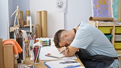 A young hispanic man with a beard appears tired, leaning on a desk full of art supplies in a creative studio.