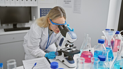 A focused woman scientist using a microscope in a laboratory surrounded by chemical flasks and...
