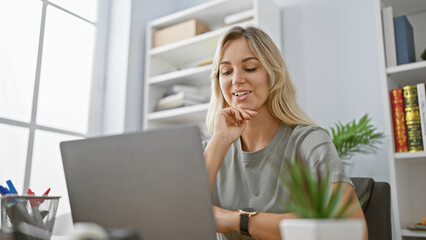 Smiling blonde woman using laptop in modern office interior, depicting professional, casual, entrepreneur, technology, career, employee, indoors.