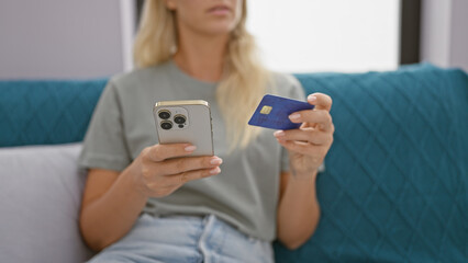 A young blonde woman holding a credit card and smartphone indoors, depicting online shopping or banking at home.