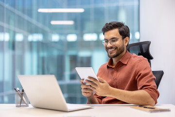 Professional sitting at office desk using tablet while working on laptop. Modern corporate environment with large windows. Concept of productivity, technology, and business success.