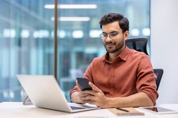 Young professional with glasses working at desk in modern office using smartphone and laptop, displaying productivity, technology, and communication. Image portrays a comfortable work environment with