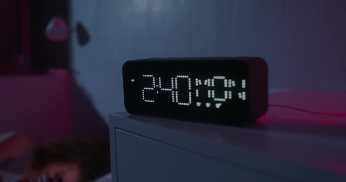 A woman uses a mobile phone lying on the bed. Dark room, home interior. The electronic clock is on the bedside table