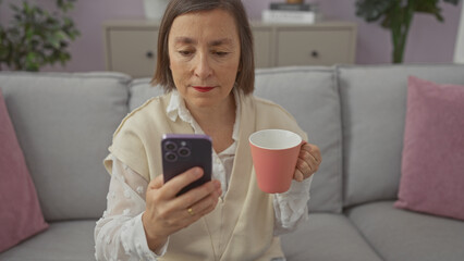 Mature woman holding mug while using smartphone on sofa indoors