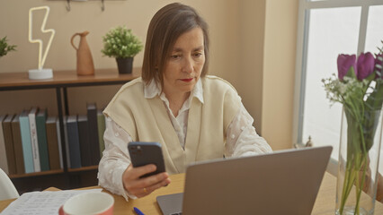 A middle-aged woman engages with a smartphone and laptop in a home office setting, exuding professionalism and focus.