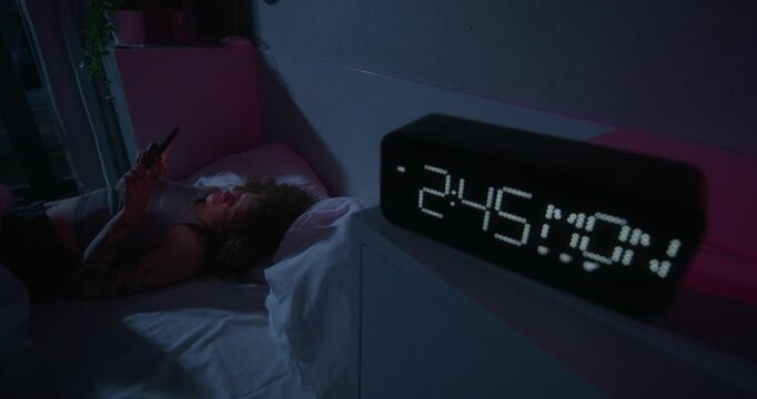A woman uses a mobile phone lying on the bed. Dark room, home interior. The electronic clock is on the bedside table