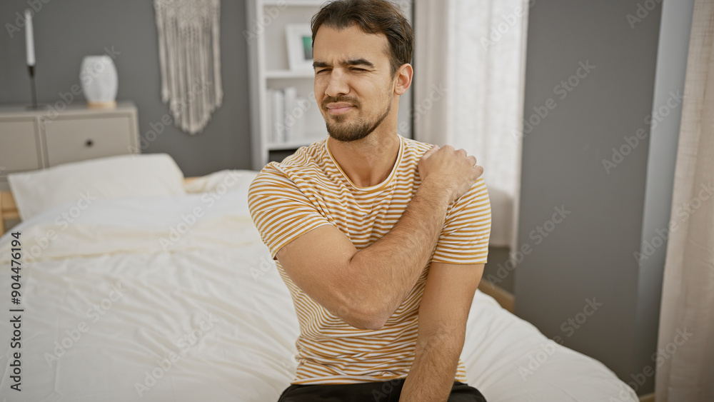 Wall mural A young hispanic man in a striped shirt shows discomfort while sitting on a bed in a well-lit bedroom, portraying a possible shoulder pain.