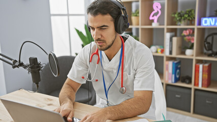 Focused hispanic man with beard in headphones and lab coat podcasting at a studio with microphone and laptop