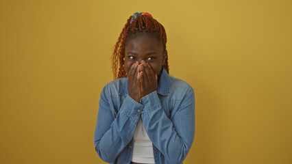 A cheerful african woman in denim over a vibrant yellow background showing a surprised expression with hands covering her mouth