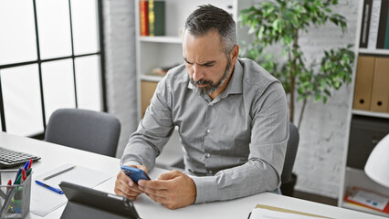 Mature hispanic man with beard and grey hair focused on smartphone in modern office.