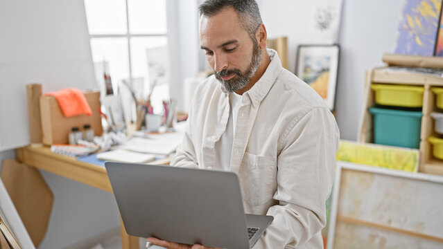 Handsome mature hispanic man with grey beard using laptop in art studio