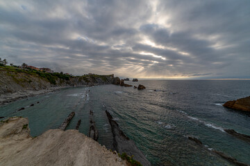 Obraz premium Beautiful sunset over Playa de la Arnía, featuring dramatic skies, rocky shoreline, and coastal homes in Cantabria, Spain. Perfect for seascape, travel, and nature themes.