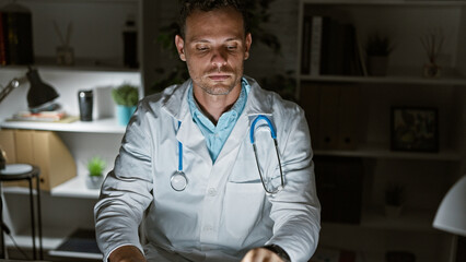 A handsome young man with a beard, dressed as a doctor, sits focused in a hospital room.