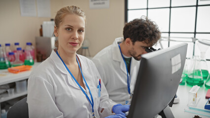 A woman and man in lab coats work together indoors, surrounded by chemistry equipment, representing a professional team in a laboratory.