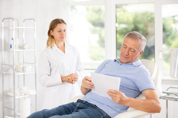 Obraz premium Old male patient lying on medical chair signing papers while talking with woman doctor