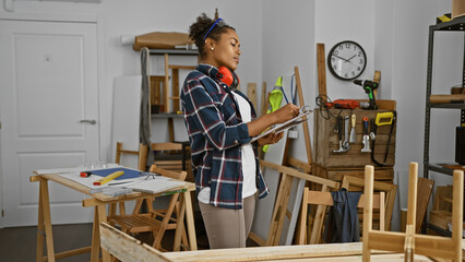 A young woman with curly hair wearing headphones takes notes in a carpentry workshop indoors.