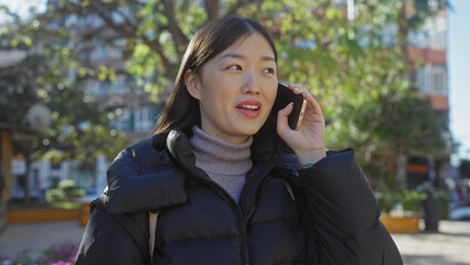 An asian woman talks on a smartphone in a sunlit urban park, exuding a casual and relaxed demeanor.