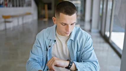 A young man checks his watch in a bright university hallway, capturing a moment of modern academic life.