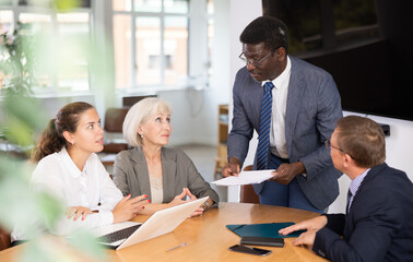 Fototapeta premium In conference room, African American man head of department speaks at meeting and reports unpleasant news to frowning colleagues. Secretary records leading moments of conversation in laptop