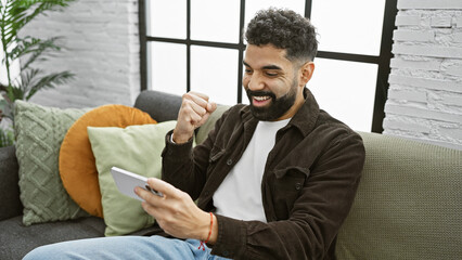 A cheerful young man with a beard celebrates a victory at home while holding a smartphone