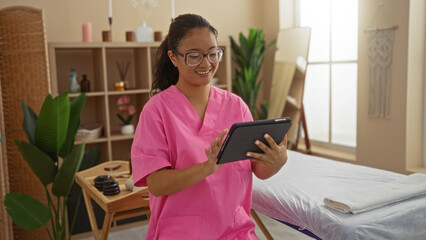 Young chinese woman in a spa setting using a tablet, dressed in pink scrubs in a peaceful wellness room with plants and a massage table, exuding a calm and professional atmosphere.