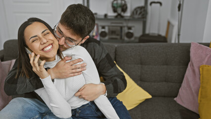 A loving couple embraces in a cozy living room, with the woman chatting on a phone and the man affectionately kissing her.