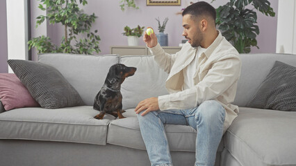 A young hispanic man plays with his teckel on a cozy couch inside a stylish apartment.